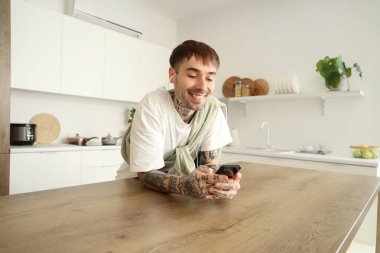 Kulaklıklı ve cep telefonlu genç dövmeli adam mutfakta müzik dinliyor.