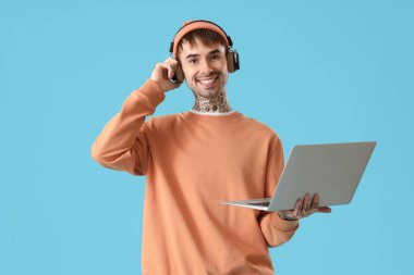 Kulaklıklı, dizüstü bilgisayarlı, mavi arka planda müzik dinleyen genç bir adam.