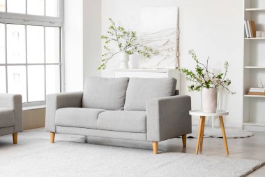 Beyaz oturma odasındaki gri kanepenin yanındaki sehpanın üzerinde çiçek açan bir vazo.