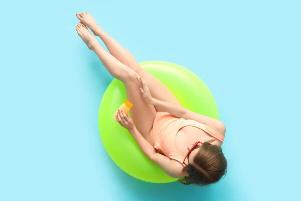 Beautiful young woman in swimsuit with bottle of sunscreen cream sitting on inflatable ring against blue background