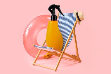 Plaj sandalyesi, şapka, şişirilebilir yüzük ve pembe arka planda bir şişe güneş kremi.