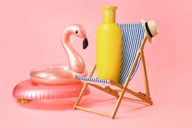 Şapkalı plaj sandalyesi, şişirilebilir halkalar ve pembe arka planda bir şişe güneş kremi.