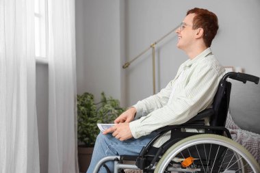 Evdeki pencerenin yanındaki tekerlekli sandalyede oturan genç adam.