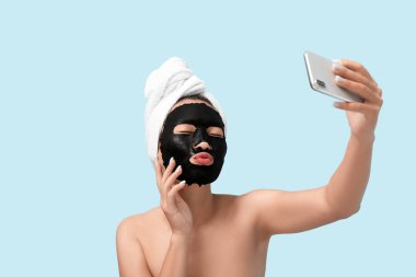 Yüzünde aktif kömür maskesi olan genç bir kadın mavi arka planda selfie çekiyor.