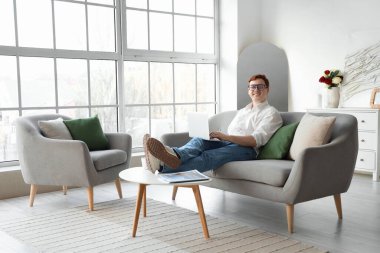 Gözlüklü genç adam evdeki gri koltukta dizüstü bilgisayar kullanıyor.