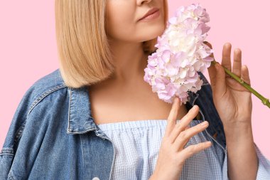 Kısa saçlı genç bir kadın pembe arka planda ortanca çiçekleri tutuyor.