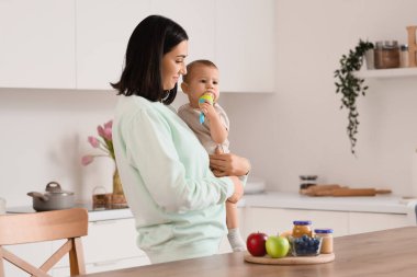 Mother giving her little baby nibbler with food in kitchen clipart