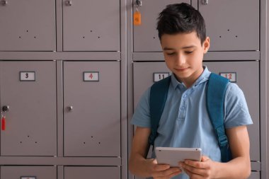 Okuldaki dolabın yanında tablet bilgisayar kullanan küçük bir çocuk.