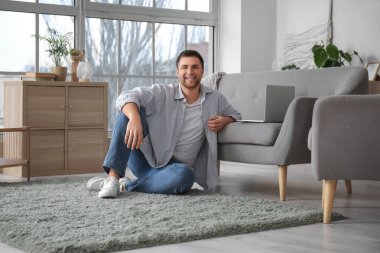 Evde dizüstü bilgisayarlı gri kanepenin yanında oturan genç adam.