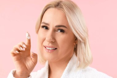 Bornozlu güzel, olgun bir kadın pembe arka plana yüz kremi sürüyor.