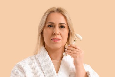 Bej arka planda yüz masajı aleti olan bornozlu güzel, olgun bir kadın.