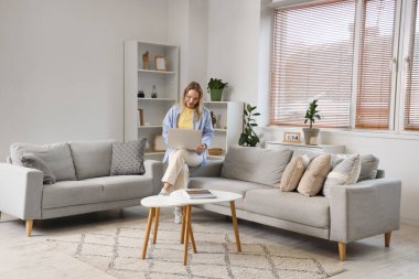 Genç bir kadın dizüstü bilgisayar kullanıyor ve oturma odasında koltukta oturuyor.