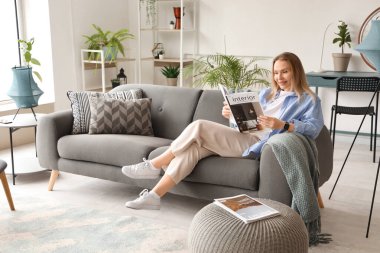 Kanepede dinlenen ve beyaz oturma odasında dergi okuyan genç bir kadın.