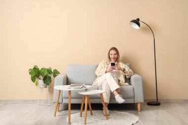 Bej oturma odasındaki koltukta oturan ve cep telefonu kullanan genç bir kadın.