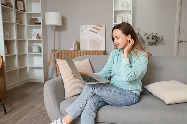 stock image Young woman using laptop on grey couch in living room