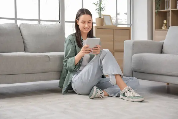 stock image Pretty young woman with tablet computer sitting on floor near grey sofa in living room