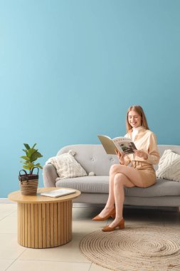 Genç ve güzel bir kadın, mavi oturma odasındaki kanepede dergi okuyor.