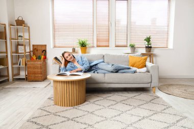 Genç, güzel bir kadın kanepede uzanıyor ve oturma odasında dergi okuyor.