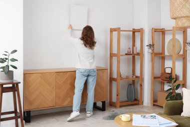 Genç bir kadın şık bir odada beyaz duvara resim asıyor.