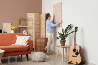 Genç, güzel bir kadın bej oturma odasındaki beyaz duvara resim asıyor.