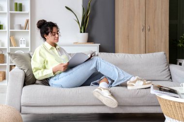 Young African-American woman reading magazine on sofa at home