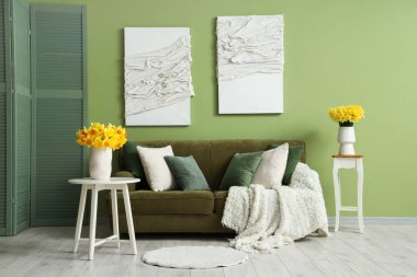 Oturma odasının içinde yeşil kanepe ve masalarda nergis var.