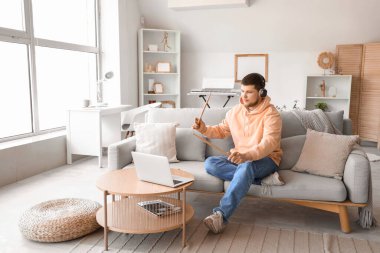 Kulaklıklı genç sakallı bir adam elinde bagetleriyle evdeki kanepede müzik dinliyor.