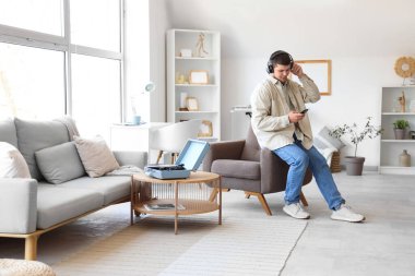 Kulaklıklı, cep telefonlu genç sakallı adam evde müzik dinliyor.