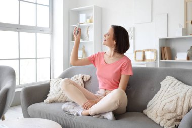 Oturma odasındaki kanepede oturan klimalı uzaktan kumandalı gülümseyen genç kadın.