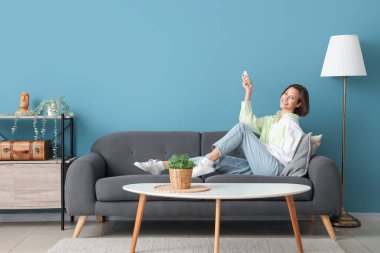 Mavi duvarın yanındaki kanepede duran klimalı uzaktan kumandalı gülümseyen genç kadın.