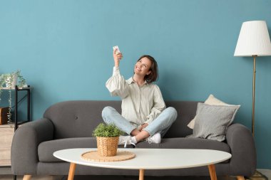 Mavi duvarın yanındaki kanepede oturan klimalı uzaktan kumandalı gülümseyen genç kadın.