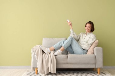 Yeşil duvarın yanındaki kanepede duran klimalı uzaktan kumandalı gülümseyen genç kadın.