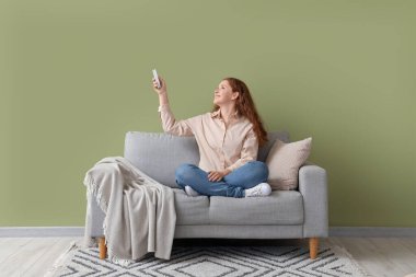 Yeşil duvarın yanındaki kanepede oturan klimalı uzaktan kumandalı gülümseyen genç kadın.