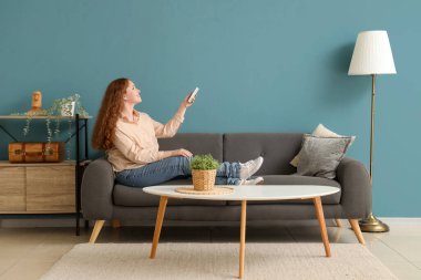 Mavi duvarın yanındaki kanepede oturan klimalı uzaktan kumandalı gülümseyen genç kadın.