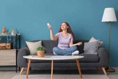 Mavi duvarın yanındaki kanepede oturan klimalı uzaktan kumandalı gülümseyen genç kadın.