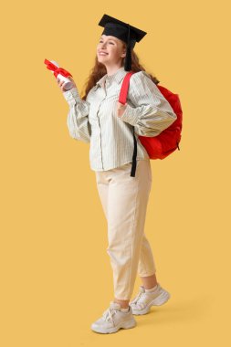 Mezuniyet şapkalı mutlu bayan öğrenci sırt çantası ve sarı arka planda diploması var.