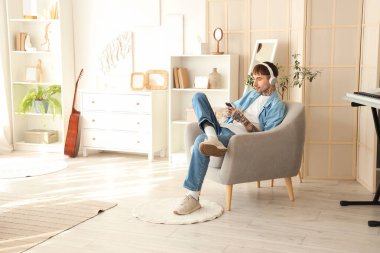 Kulaklıklı, cep telefonlu, evdeki koltukta müzik dinleyen genç dövmeli adam.