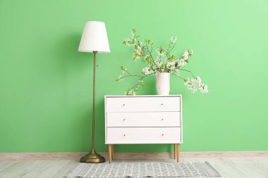 Yeşil odada, lambanın yanındaki beyaz çekmecelerin üzerinde çiçek açan bir vazo.