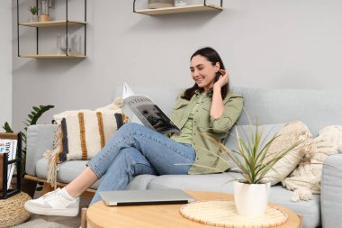 Genç bir kadın, evdeki duvarın yanındaki gri koltukta dergi okuyor.