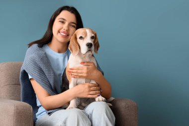 Evdeki mavi duvarın yanındaki gri koltukta oturan sevimli köpekli genç kadın.