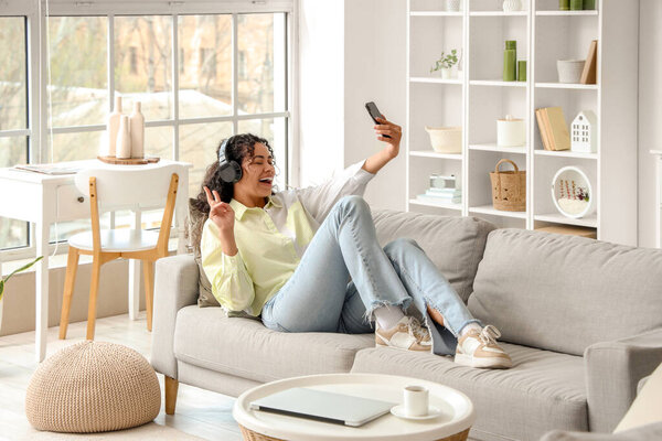 Young African-American woman in headphones with mobile phone taking selfie on sofa at home