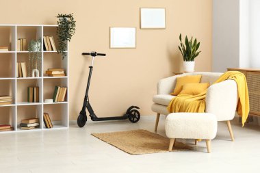 Oturma odasındaki bej duvarın yanında elektrikli scooter, koltuk ve kitaplık.