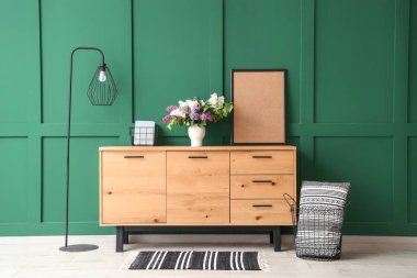 Ahşap dolapta çiçekli vazo, lamba ve yeşil duvarın yanındaki oda.