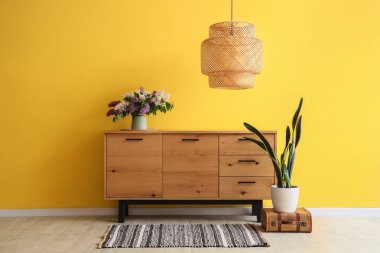 Ahşap dolapta çiçekli vazo, lamba ve oda sarı duvarın yanında bitkiler.