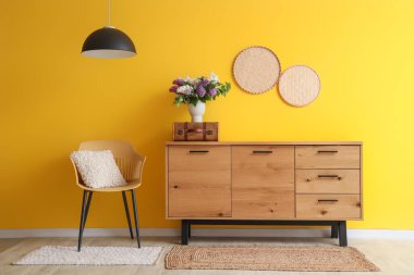 Ahşap dolapta çiçekli vazo, oda sarı duvarın yanında lamba ve sandalye.