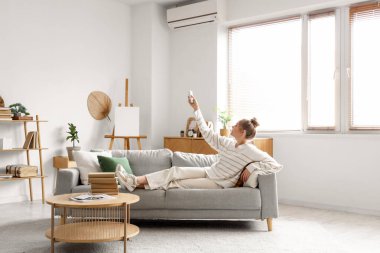 Genç bir kadın uzaktan kumandayla saç kremini açıyor ve oturma odasında kanepede uzanıyor.