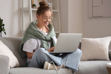 Gözlüklü genç kadın kanepede oturuyor ve oturma odasında dizüstü bilgisayarla sohbet ediyor.