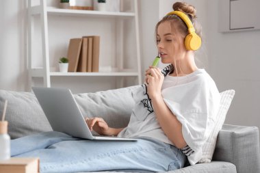 Kulaklıklı, tek kullanımlık sigara içen ve evdeki kanepede müzik dinleyen genç bir kadın.