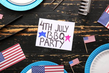 Koyu ahşap arka planda Bağımsızlık Günü kutlamaları için 4 Temmuz barbekü partisi, yemek takımı ve ABD bayrakları içeren tebrik kartı.