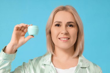 Mavi arka planda bir kavanoz yüz kremi olan güzel, olgun bir kadın.